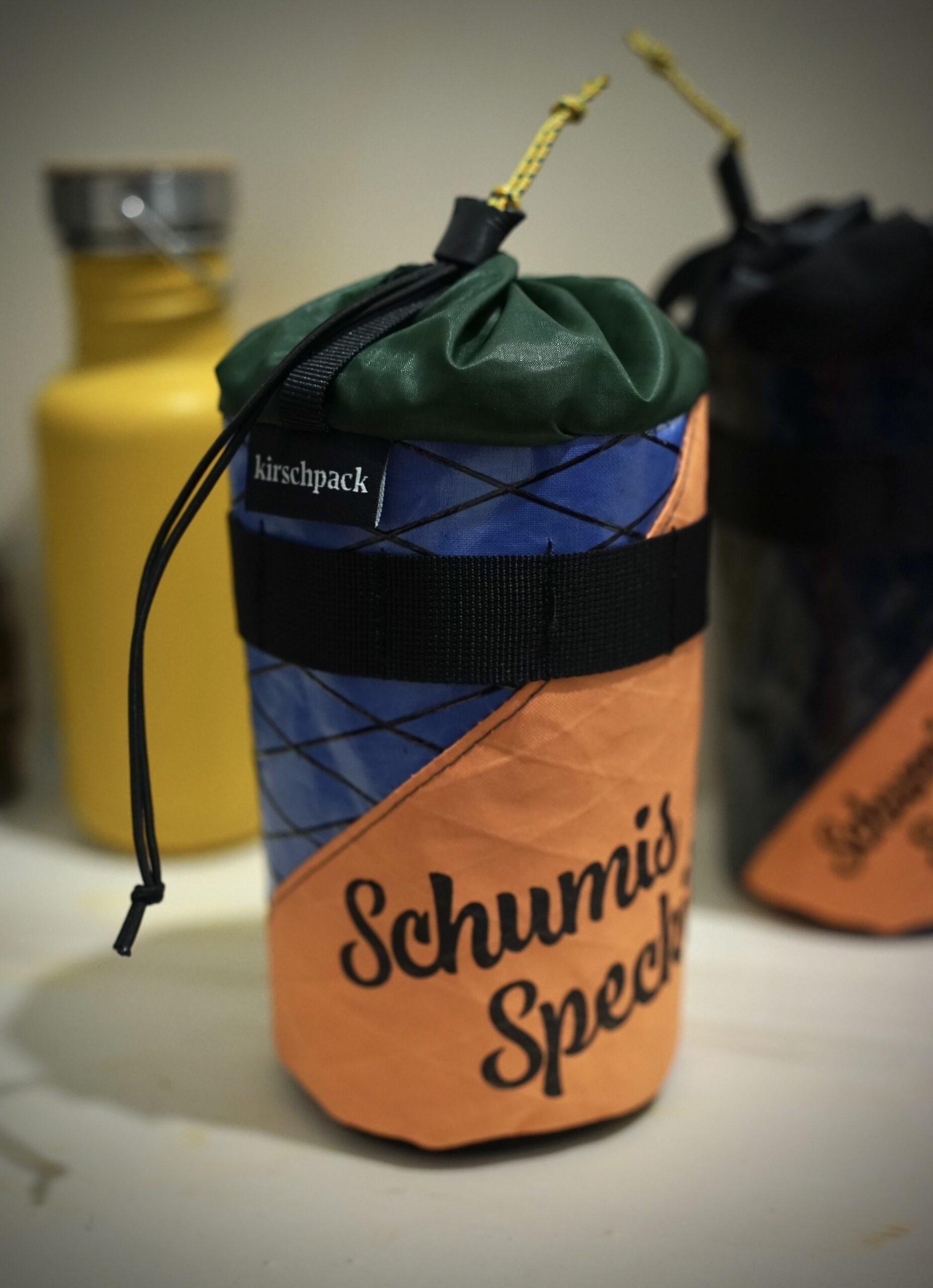 Snackbag von kirschpack in Orange, Blau und Grün mit Aufdruck »Schumis Speckilon« und Kordelzug neben einer Trinkflasche